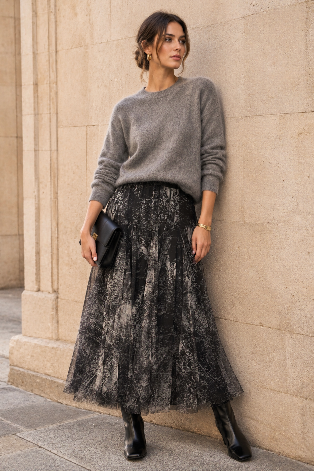 A woman stands against a beige stone wall wearing a gray knit sweater, a long black lace tulle midi skirt, black ankle boots, and holding a black clutch.