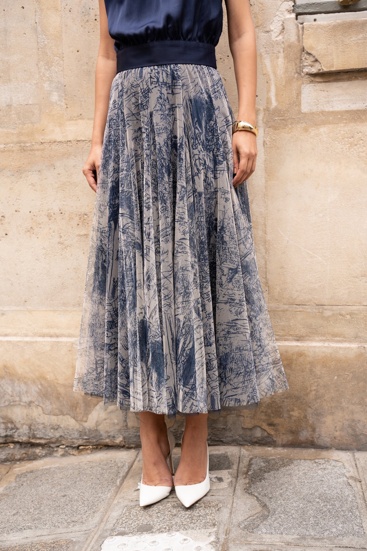 Person wearing a patterned skirt and white shoes against a stone wall.