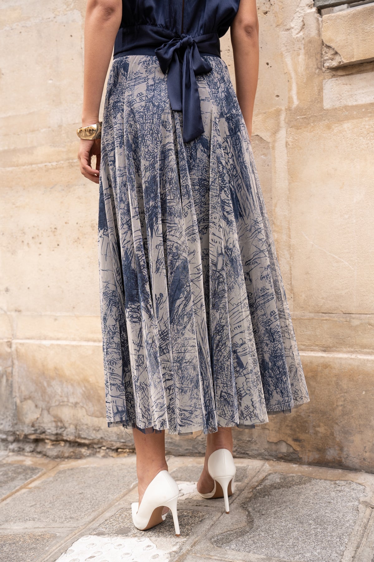 Person wearing a blue patterned skirt and white shoes against a stone wall.