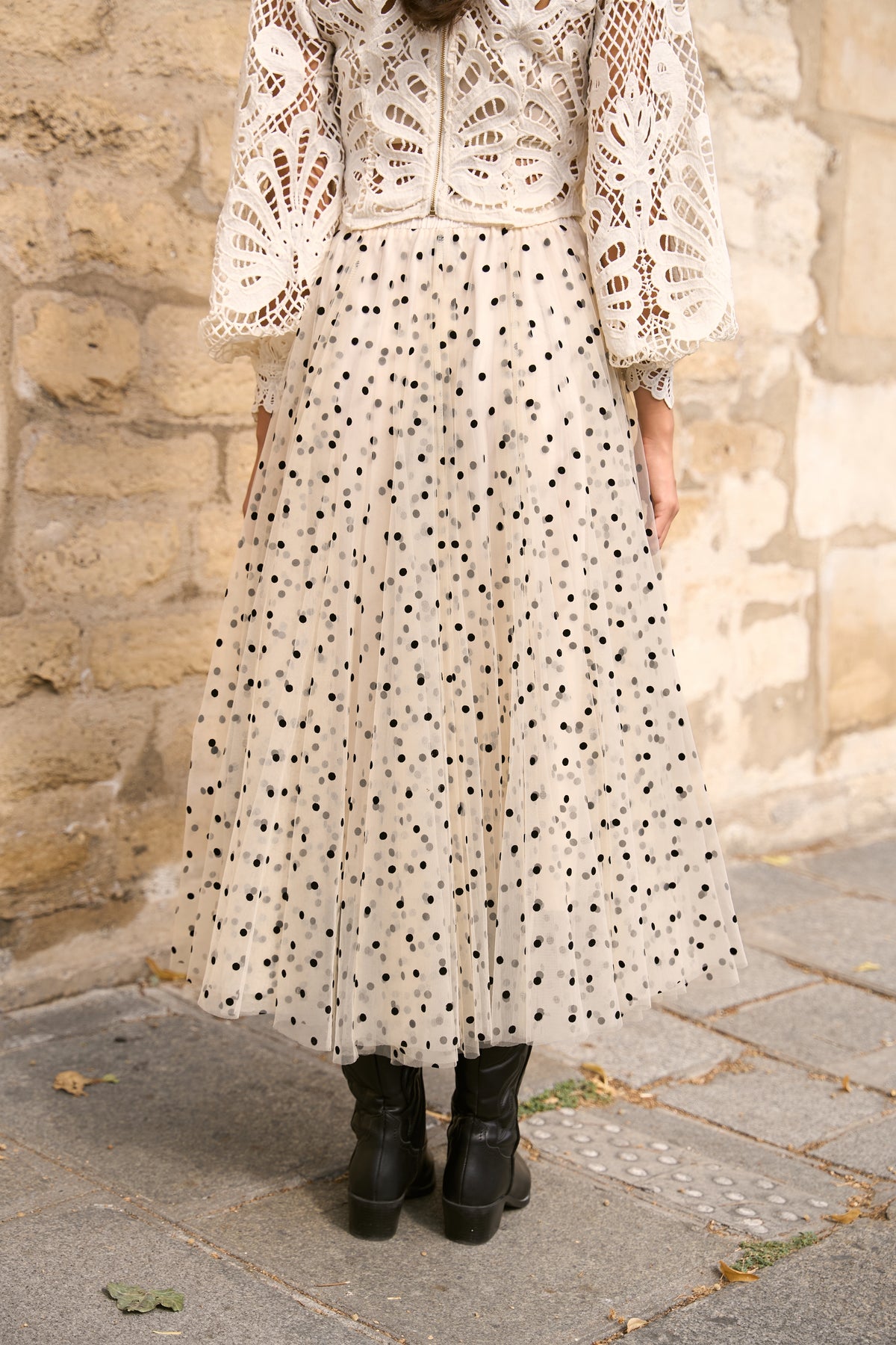 A person wearing a cream-colored bohemian-style maxi dress with black polka dots and a matching lace crochet cardigan, standing outdoors against a stone wall.