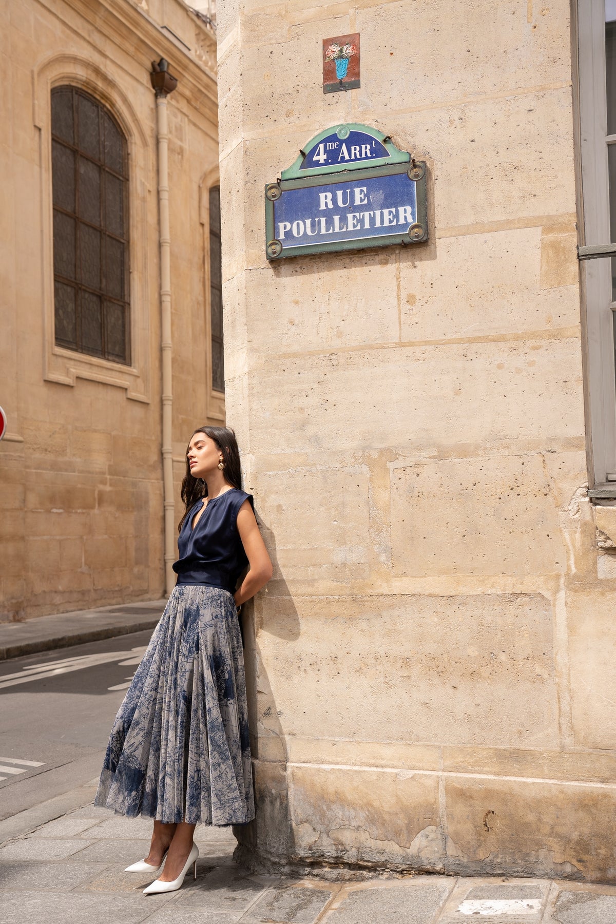 A woman wearing a navy blue satin top and a blue-and-white patterned pleated midi skirt leans against a beige stone building wall next to a blue 'Rue Poulletier' street sign.