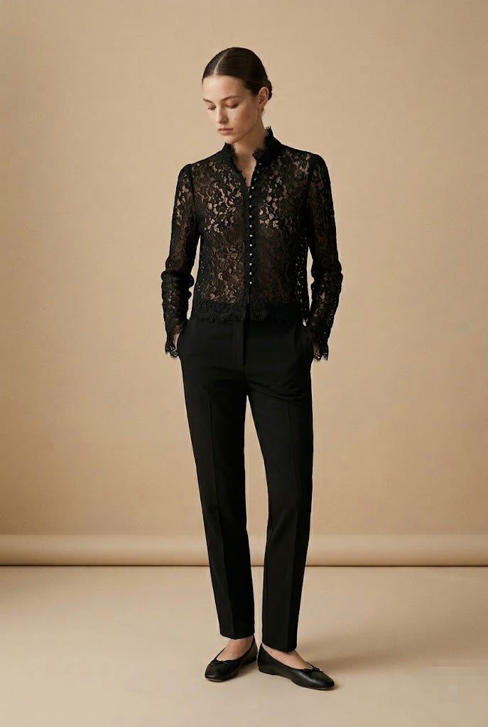 A woman stands in a studio wearing a black lace long-sleeve button-front blouse with black trousers and black flats, against a warm beige backdrop.