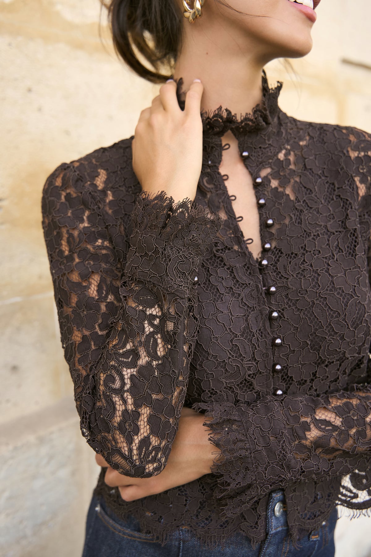 A close-up of a woman wearing a dark brown lace long-sleeve blouse with a ruffled mock neck and a front button placket, paired with blue jeans against a light stone wall.