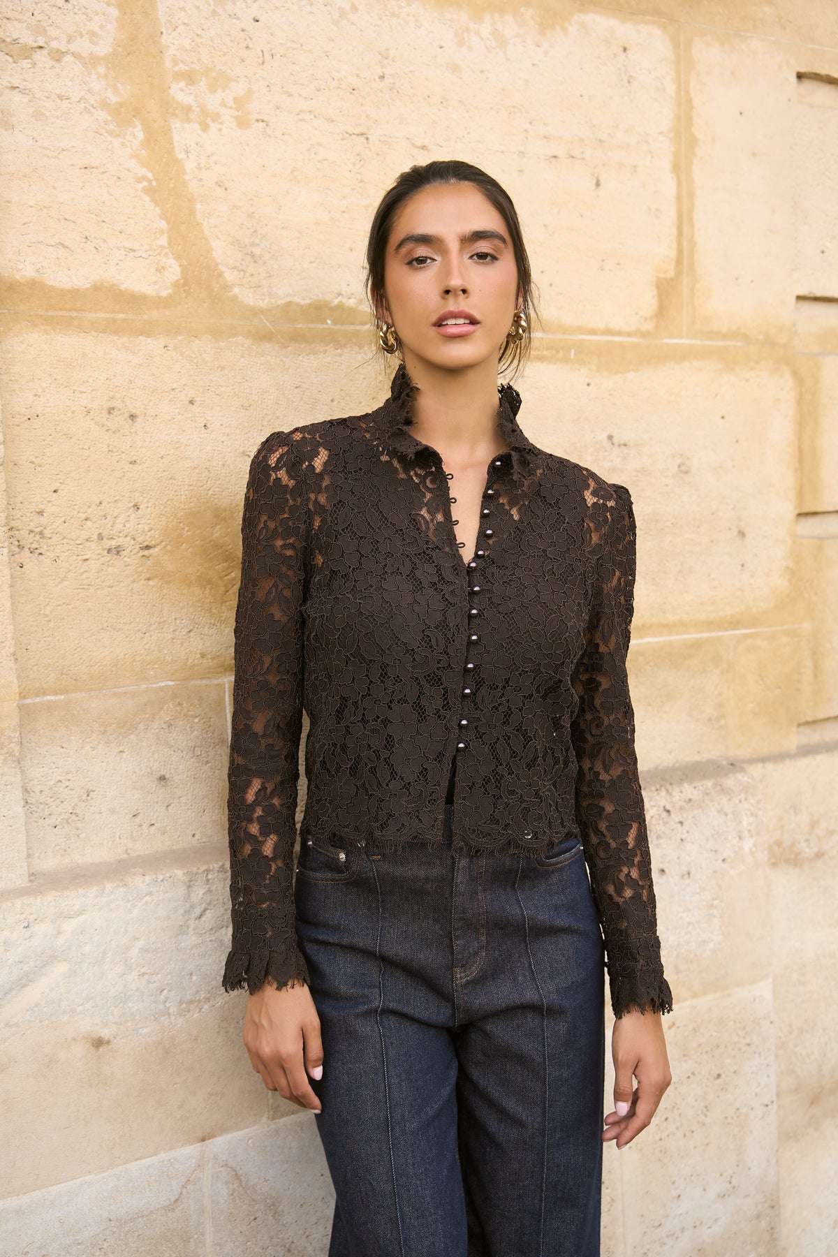 A woman wearing a dark brown lace long-sleeve button-up blouse and dark blue jeans, standing against a beige stone wall.