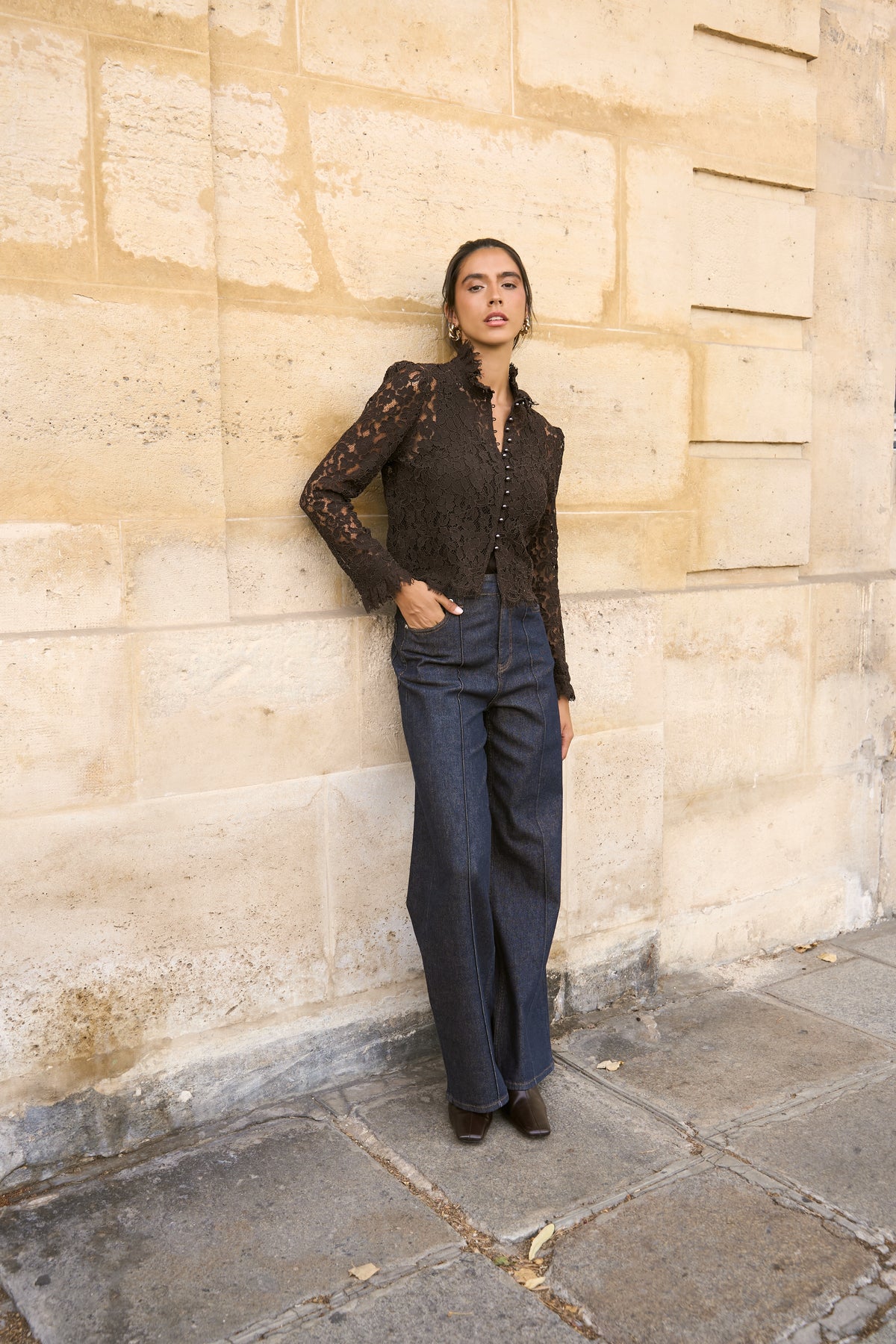 A woman stands against a beige stone wall wearing a dark brown lace long-sleeve top and dark blue wide-leg jeans.