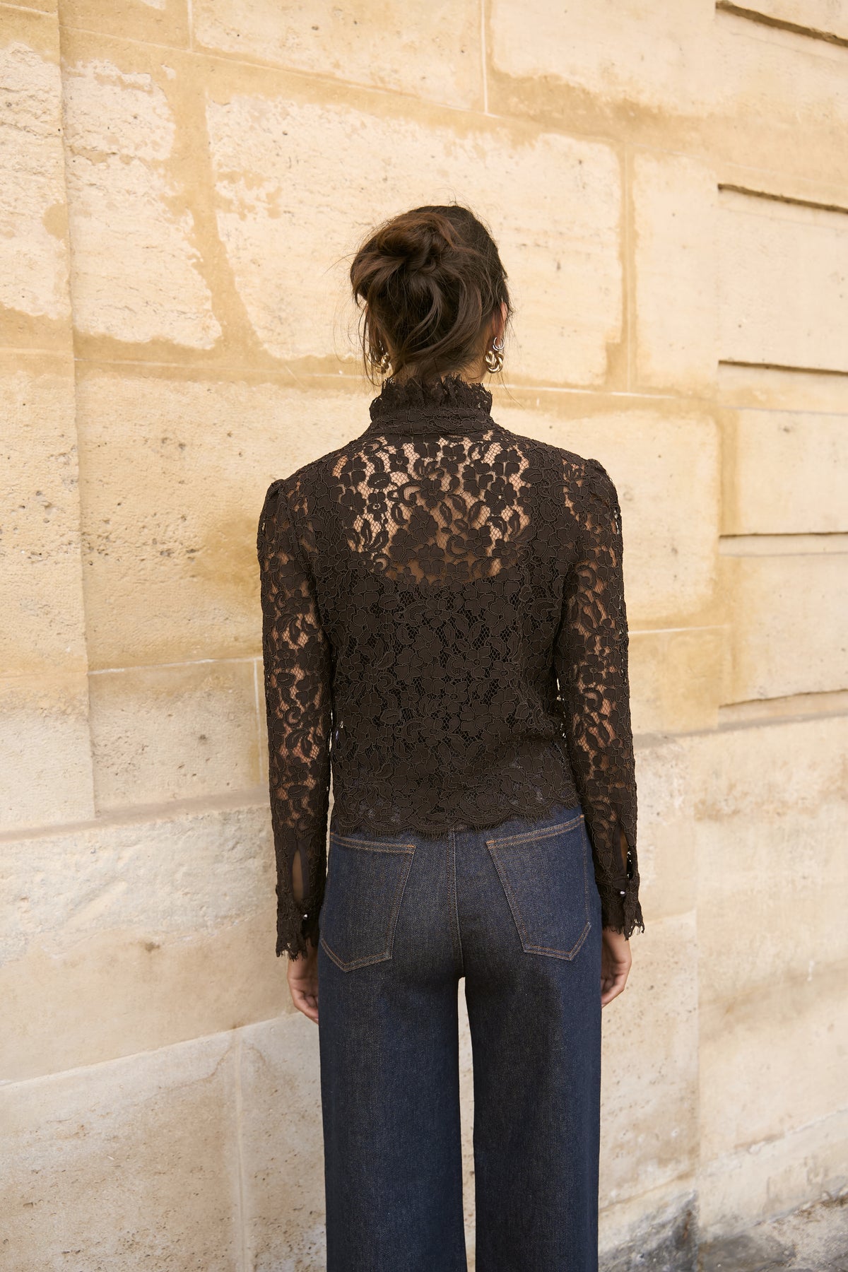 A woman viewed from behind wearing a black lace mock-neck long-sleeve top and dark blue jeans, standing against a beige stone wall.